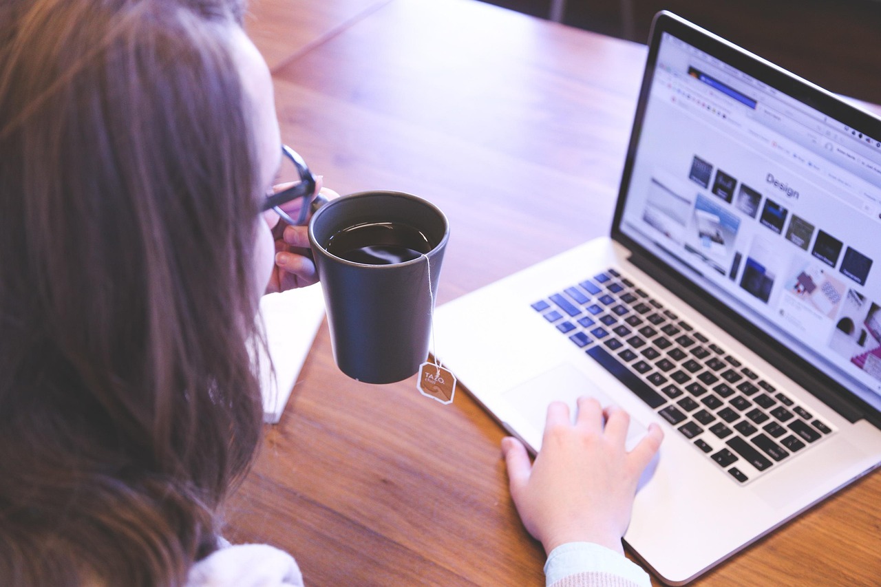 Mulher de óculos toma chá em uma caneca enquanto navega em um notebook com uma página de design de sites na tela, sobre mesa de madeira.